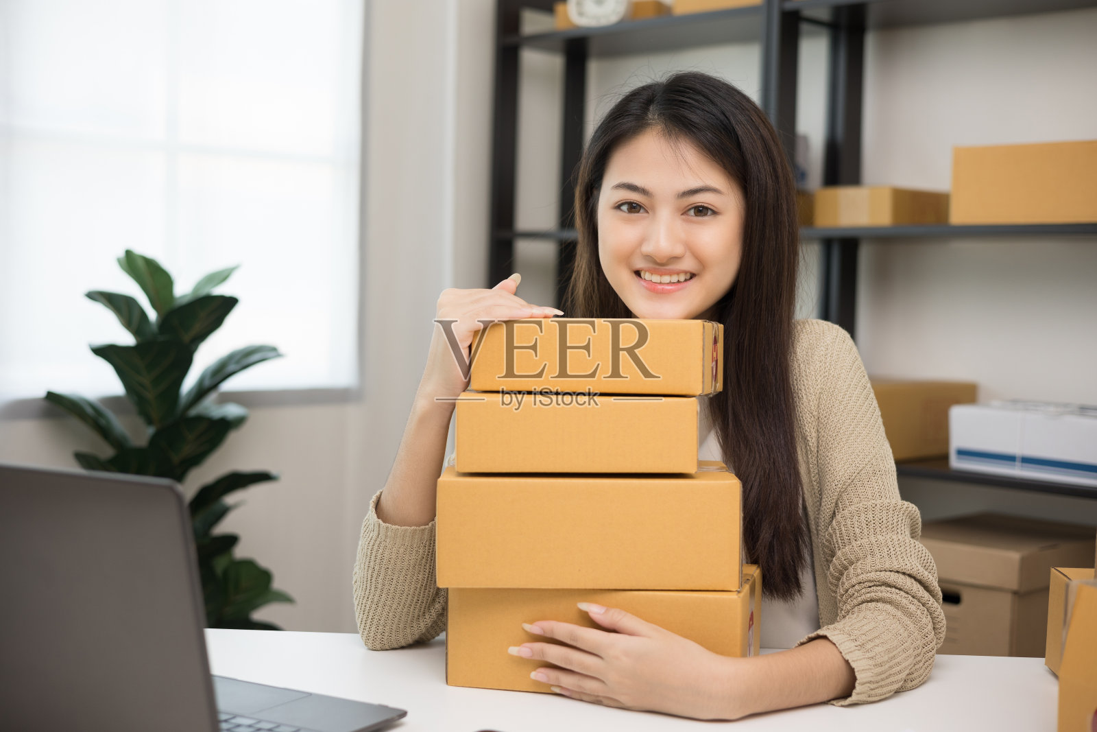 年轻美丽的亚洲企业家堆叠包裹纸板和拥抱产品和微笑的相机。快乐的商业女性与许多盒子在家里工作。小企业主创业的概念。照片摄影图片