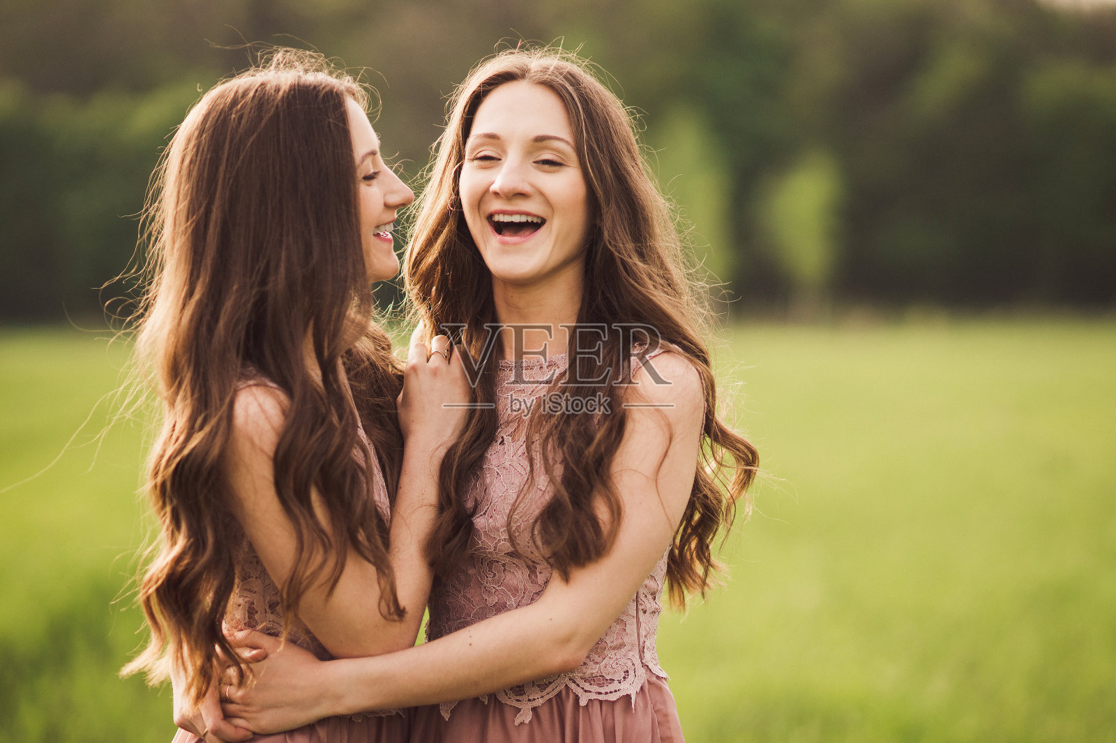 美丽快乐的双胞胎姐妹穿着长长的晚礼服，在阳光下拥抱在春天的绿色田野里。照片摄影图片