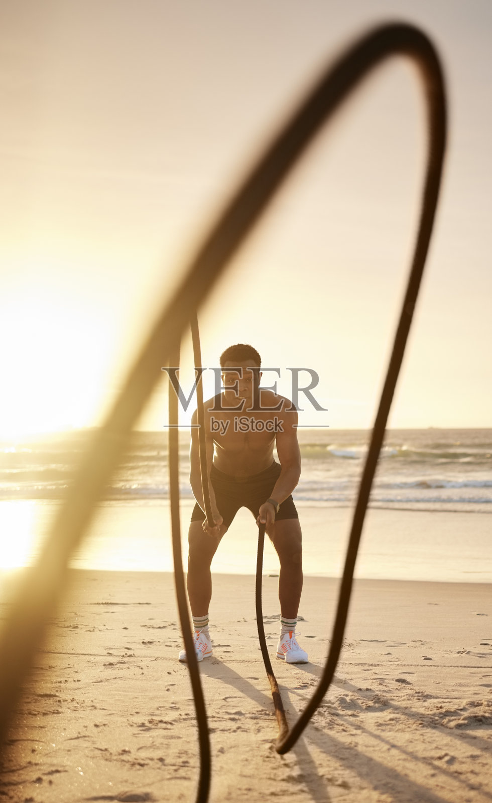 一名黑人男子在海滩上用两根战绳进行锻炼。健美的非洲裔美国男性运动员在海边锻炼。致力于健康的生活方式。日落时分在海边晨练照片摄影图片