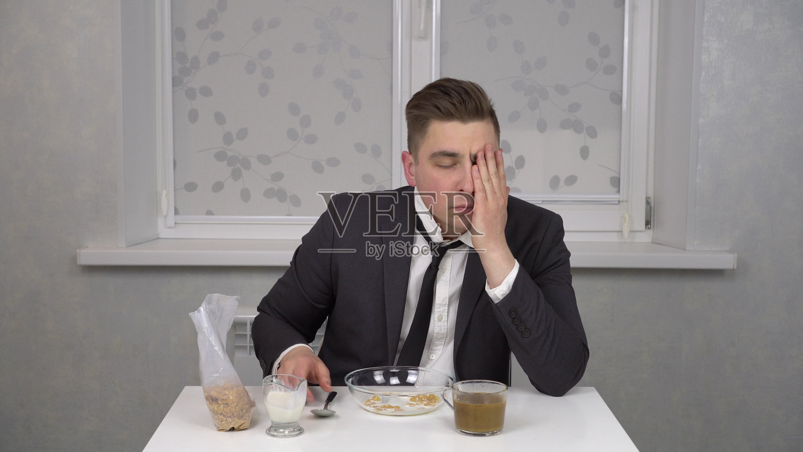 一个穿着西装的昏昏欲睡的年轻人吃麦片，喝咖啡。年轻的商人在家里的厨房里。照片摄影图片