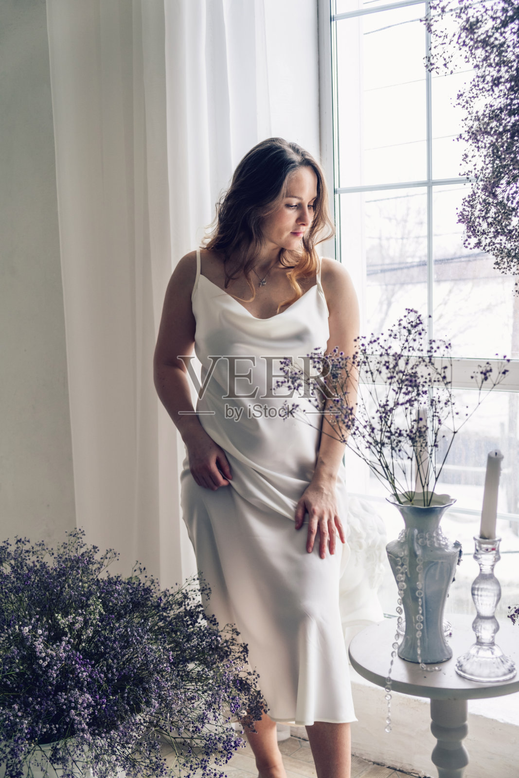 年轻美丽的长发女子穿着轻盈的丝绸裙，在装饰着2022年的gypsophila时尚色彩的房间里望向窗外照片摄影图片