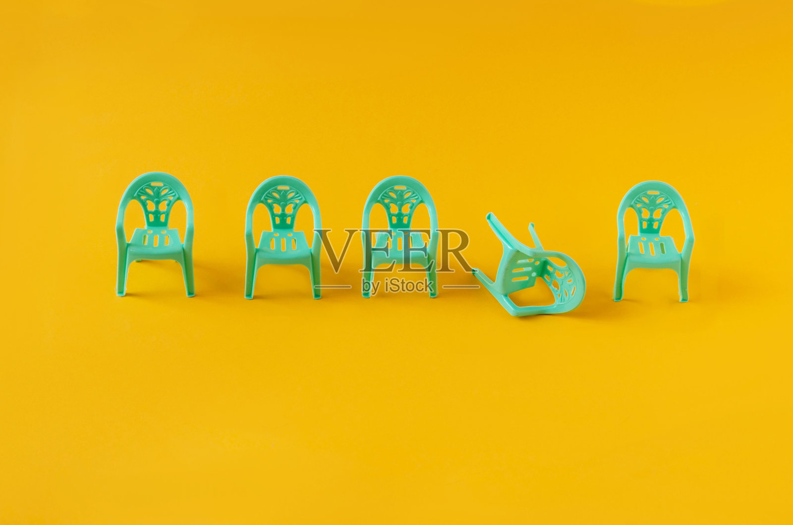 四把蓝绿塑料椅子排成一排，其中一把翻倒在黄色背景下。对于意见分歧、混乱或讨论的最小创造性概念。象征性的设计照片摄影图片