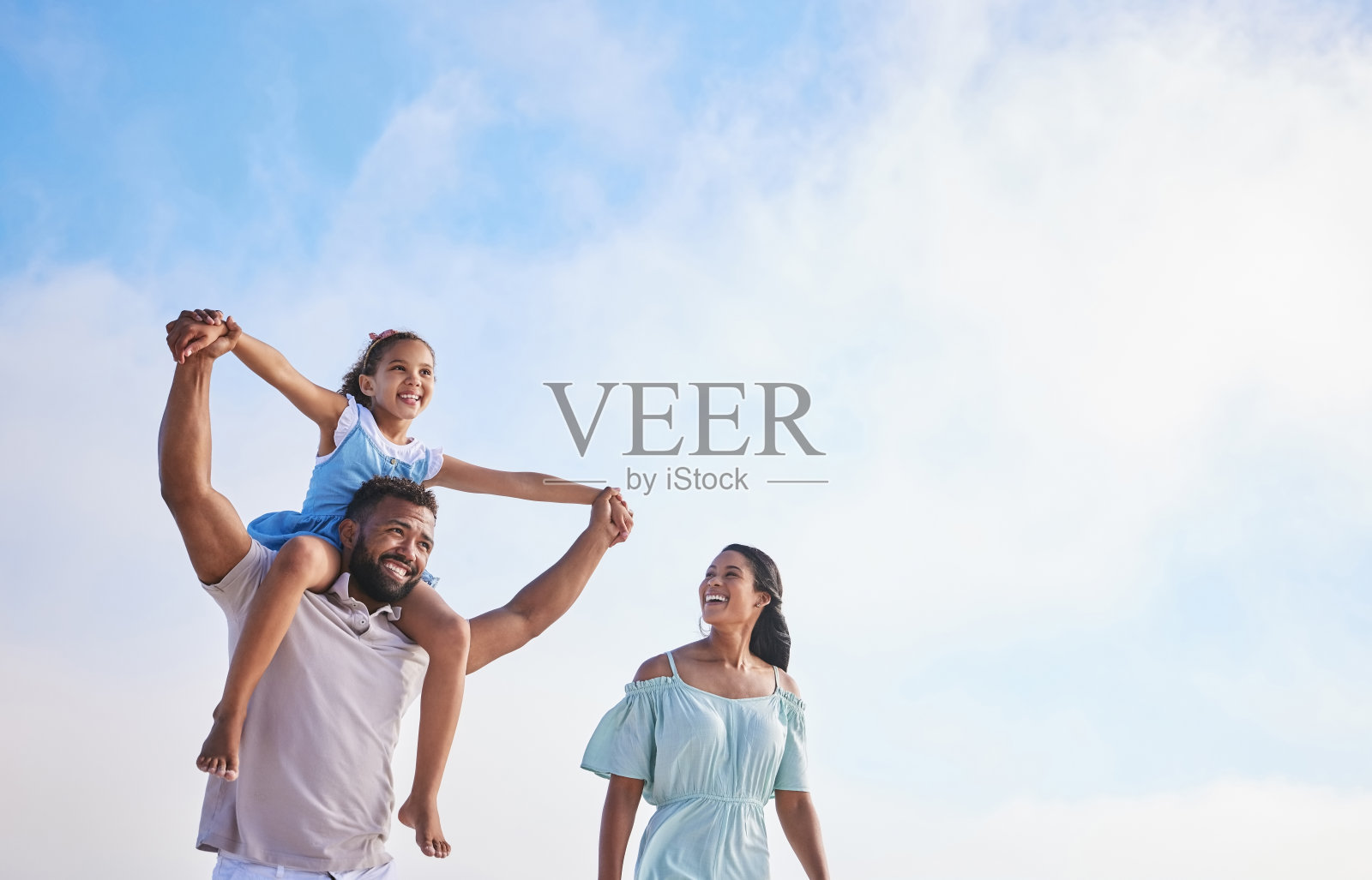 慈爱的父亲扛着女儿，和妻子在多云的天空下走在海滩上。混合种族家庭在旅行和度假时共度时光照片摄影图片