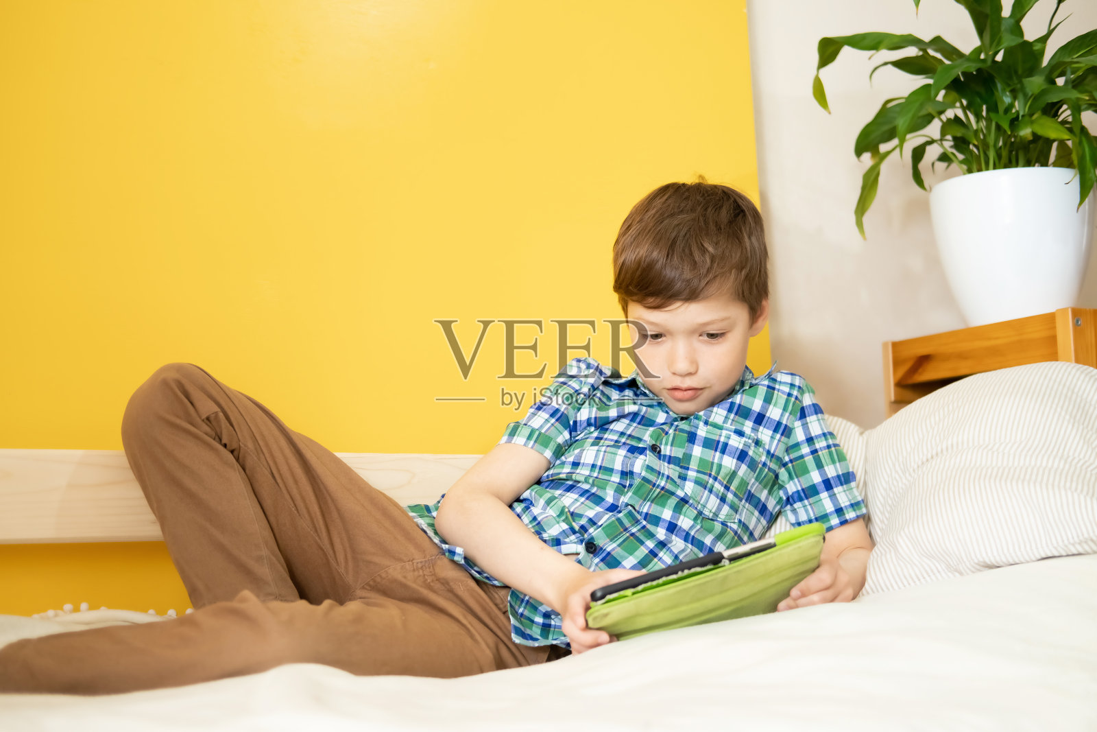 一名白人男孩躺在床上，拿着平板电脑玩网络游戏，上网，在社交网络上交流。儿童对电子产品的依赖。照片摄影图片