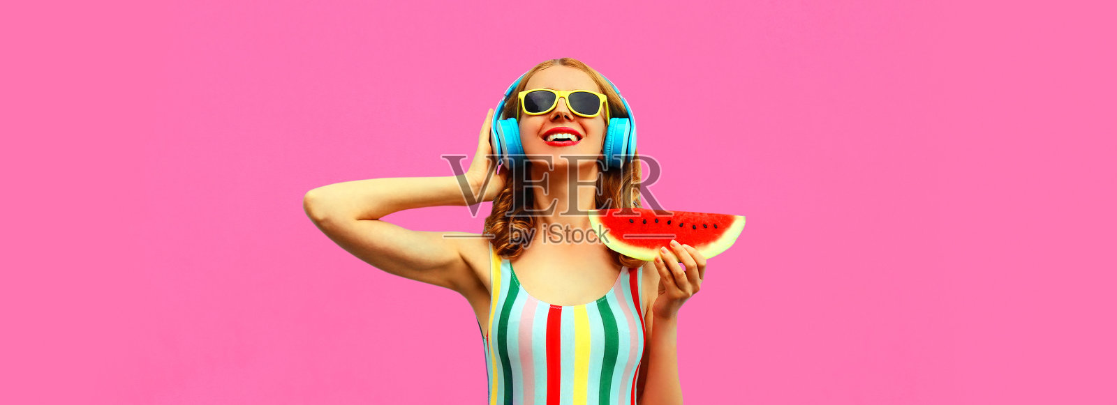 夏天彩色肖像开朗快乐微笑的年轻女子模特戴着耳机听音乐多汁的西瓜片在粉红色的背景，空白的拷贝空间的广告文字照片摄影图片