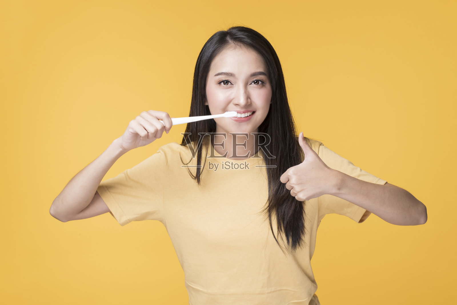 幸福的亚洲女性刷牙的肖像。快乐的亚洲女人穿着黄色t恤站在黄色的背景。照片摄影图片