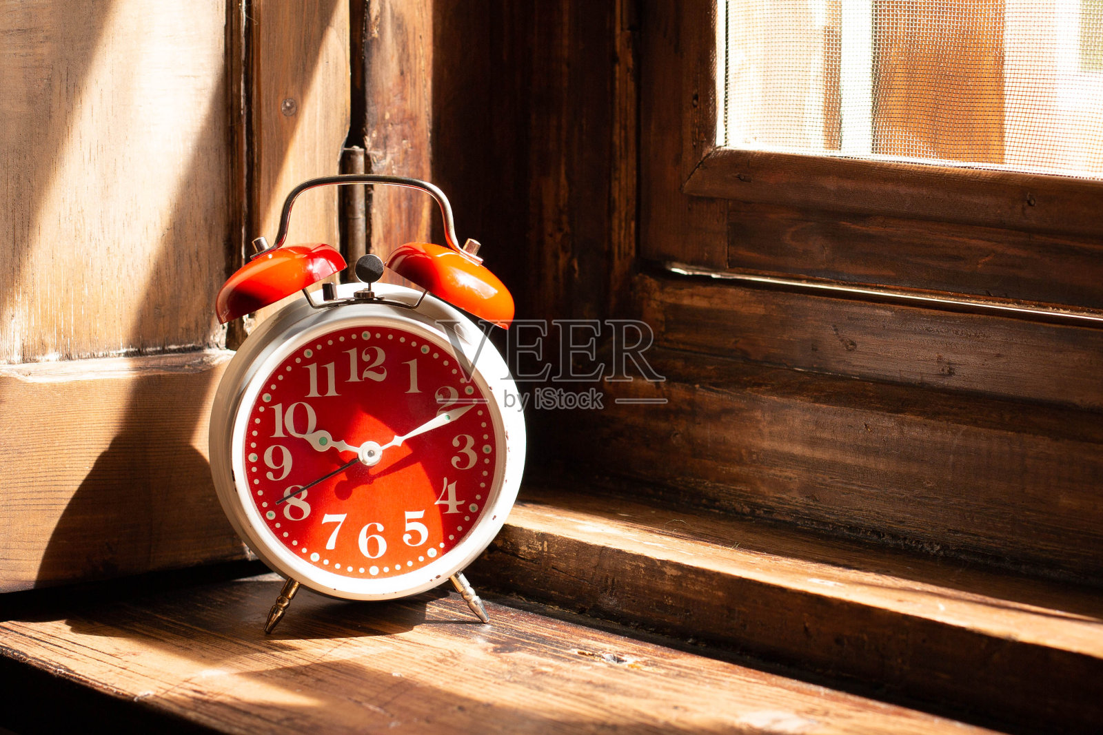 一个红色的闹钟立在木制的窗台上，被早晨的阳光照得透亮。照片摄影图片