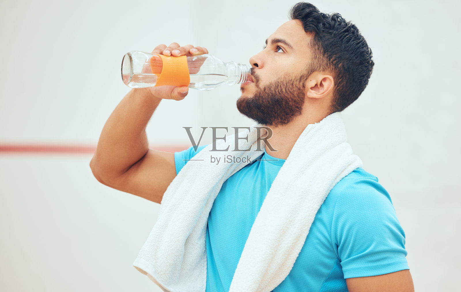 年轻的壁球运动员在打完球场比赛后独自站着喝水。适合在运动中心训练训练后休息的活跃的混合种族运动员。西班牙裔运动男子在休息照片摄影图片