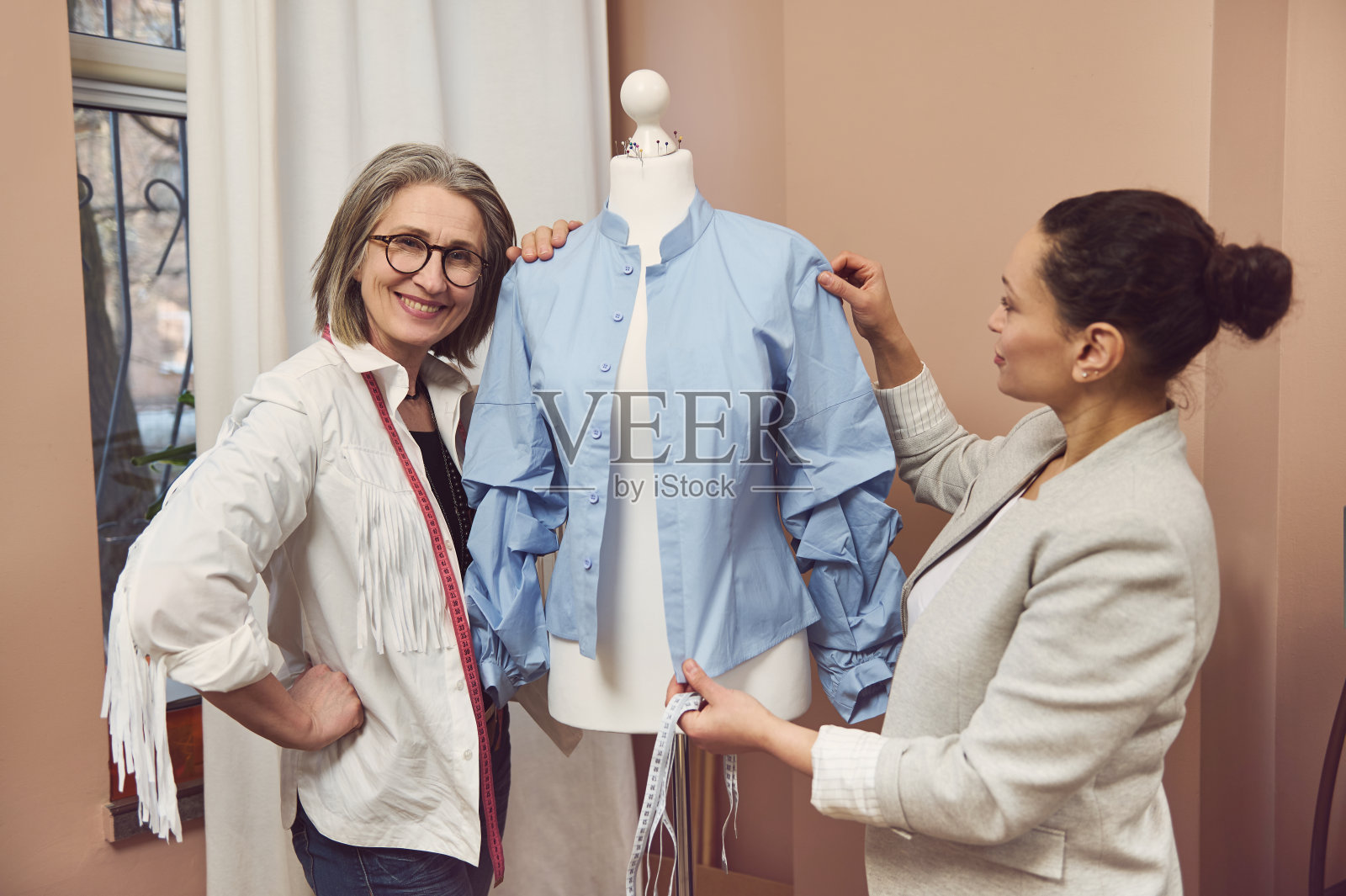 年长的时装设计师微笑地与穿着新款蓝色衬衫的人体模型合影，她的同事正在测量并改进新服装的细节照片摄影图片