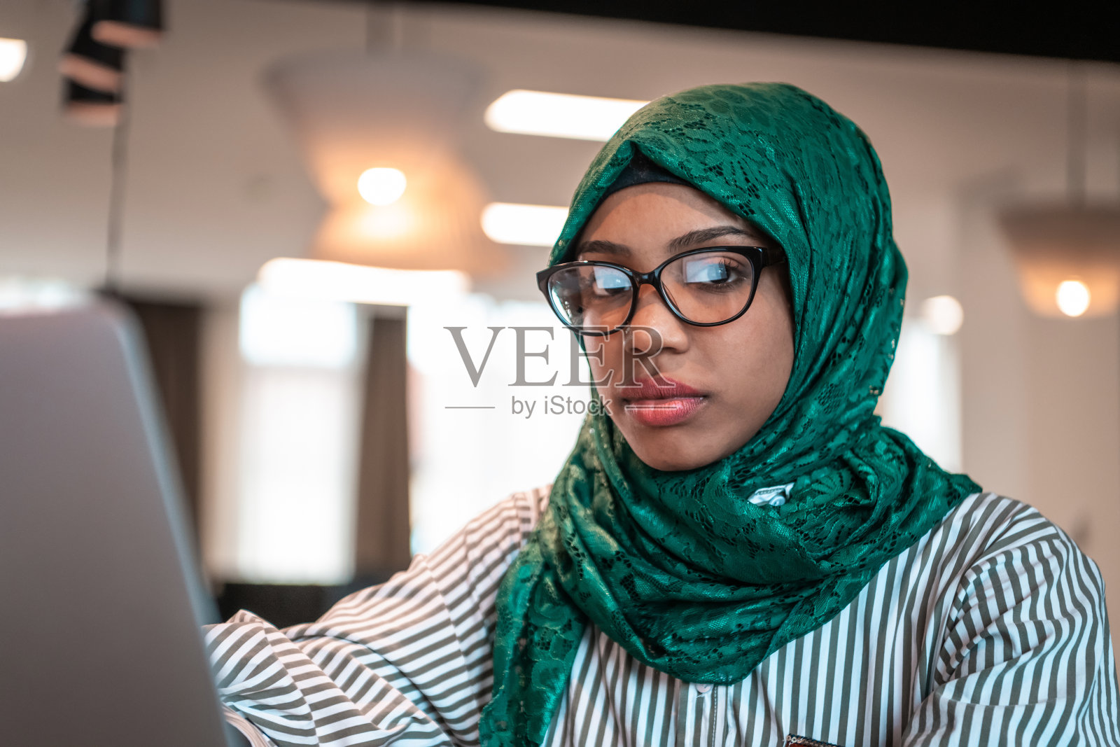 在现代开放式创业办公室的休闲区，戴着绿色头巾的商务女性使用笔记本电脑。有选择性的重点照片摄影图片