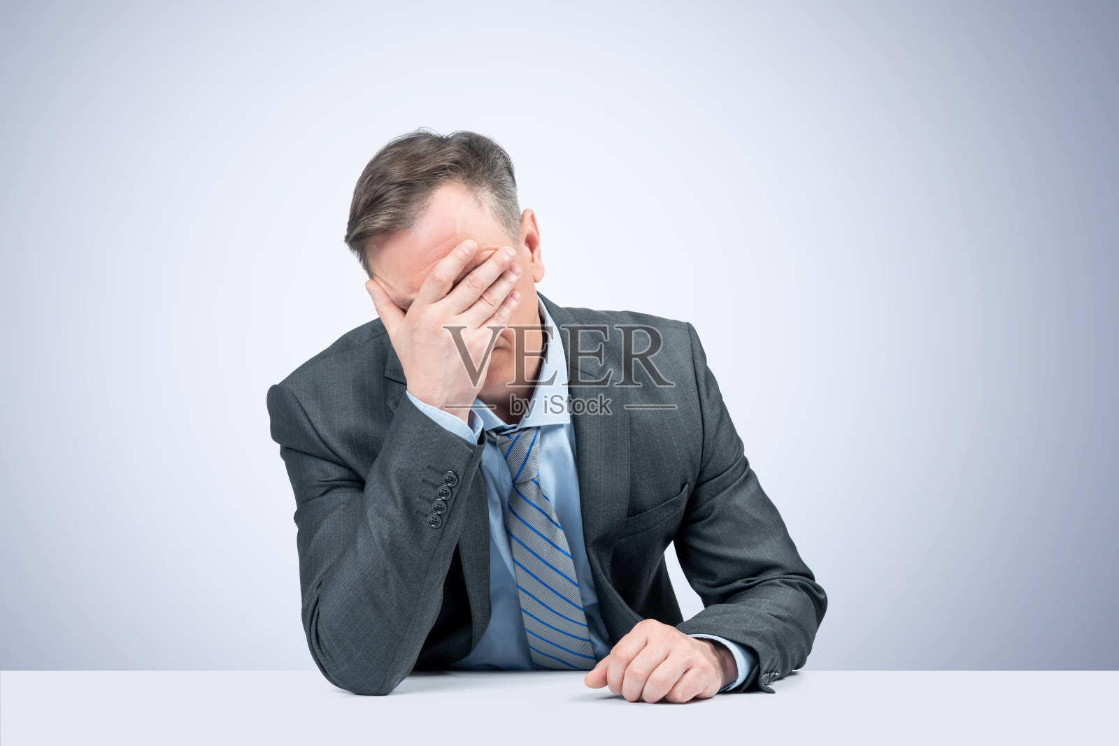 一个穿着深色夹克、衬衫和领带的男人坐在桌子旁，在浅蓝色的背景下做了一个捂脸的动作。史诗般的失败的情感。照片摄影图片