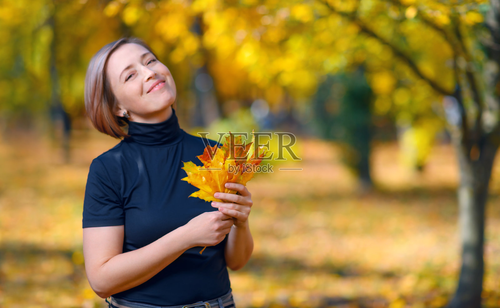 一位美丽的女子在秋天的城市公园与黄色的树叶摆姿势，捧着一束树叶，阳光明媚的日子照片摄影图片