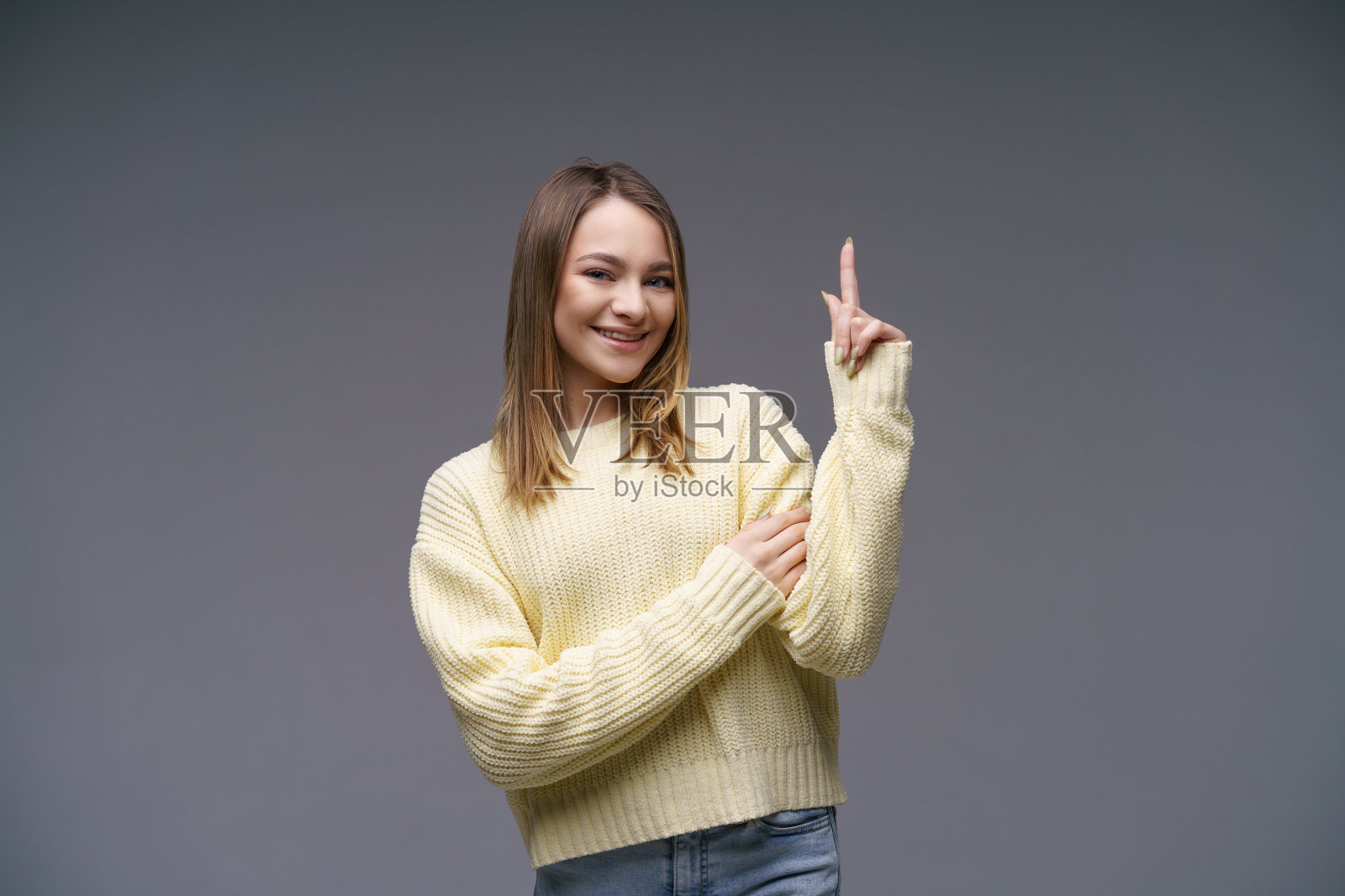 笑容满面的年轻女人在灰色背景下的黄色毛衣上竖起大拇指照片摄影图片