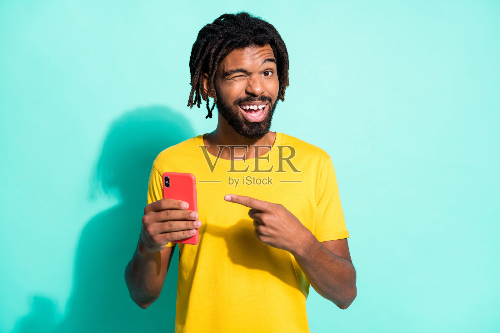 照片的年轻微笑非洲人与棕色脏辫眨眼的眼睛点手指手机孤立在蓝绿色的背景照片摄影图片