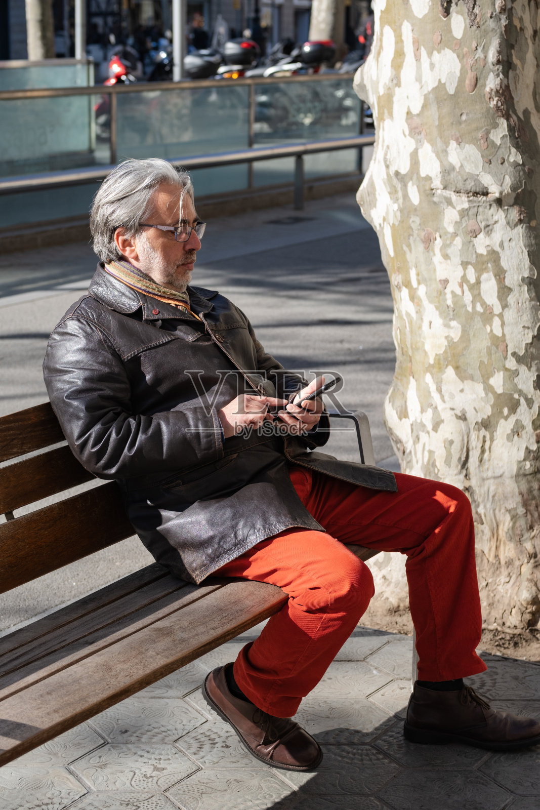 一名白发大胡子的成年男子坐在树旁的木凳上打电话照片摄影图片