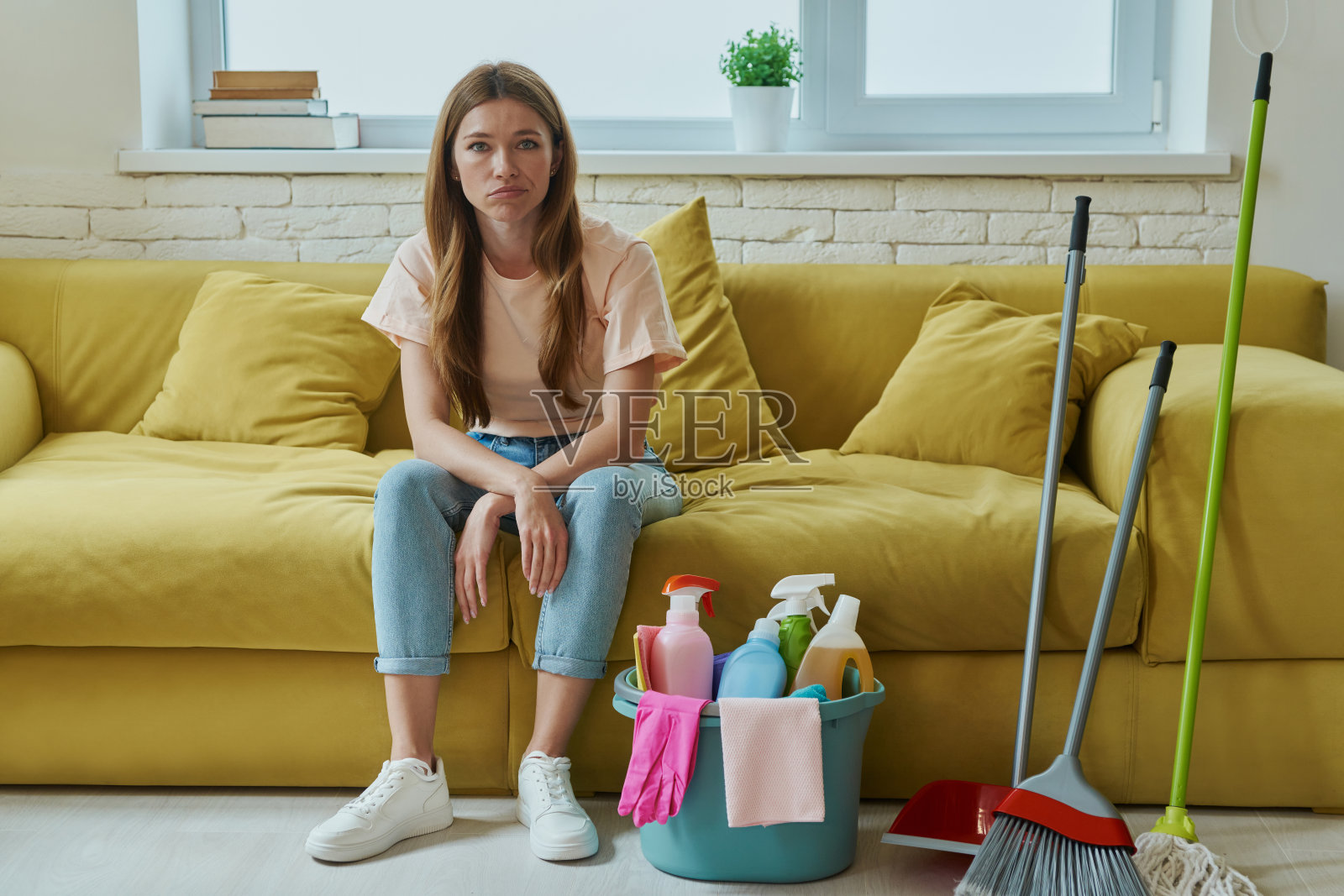 沮丧的女人坐在沙发上，手里拿着一桶清洁用品照片摄影图片