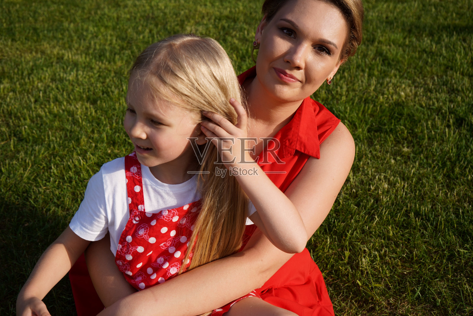 身穿红色背心和白色t恤、金发碧眼的5岁欧洲小女孩身穿红色连衣裙，坐在绿色草地上的母亲旁边照片摄影图片