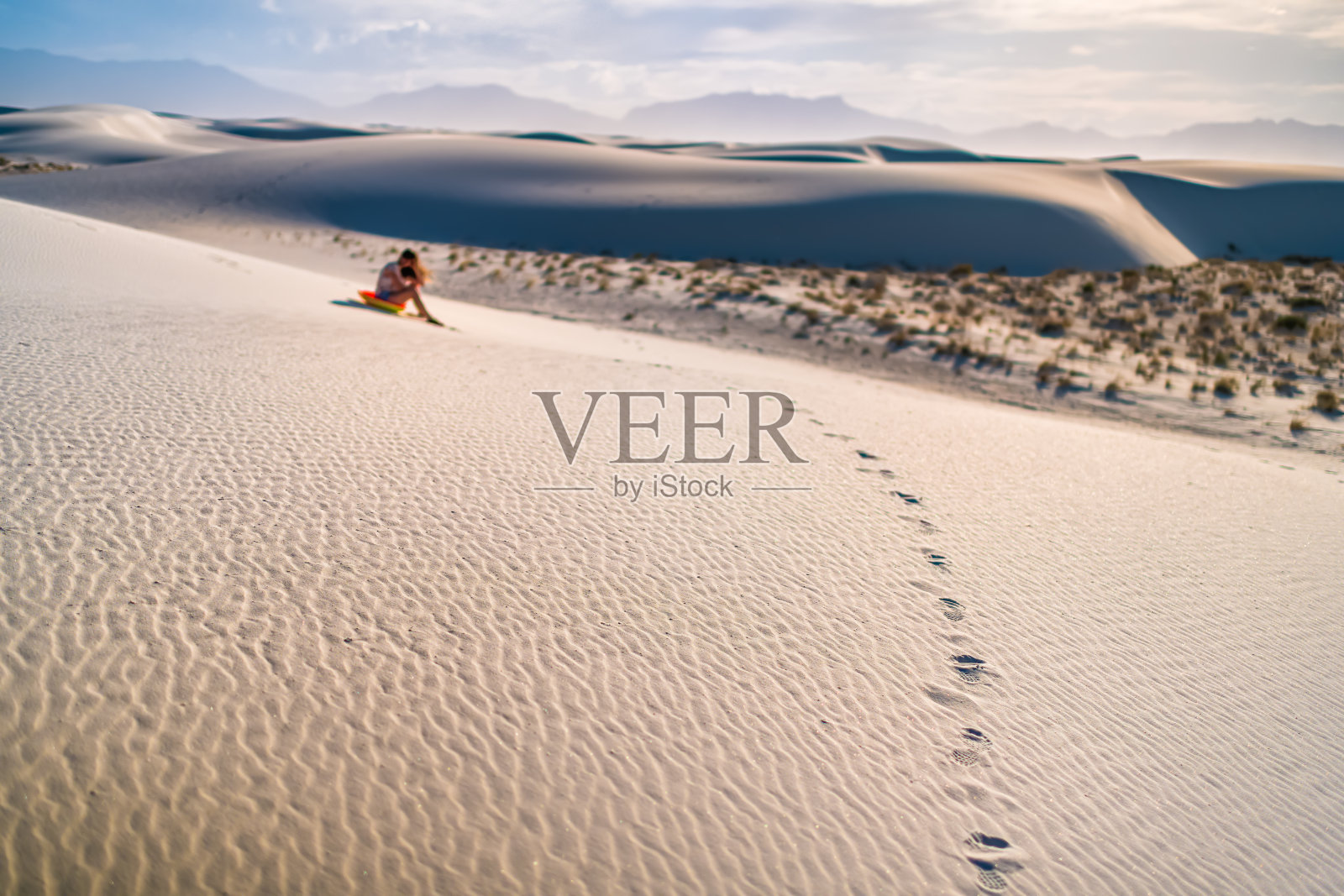 在新墨西哥州著名的白沙沙丘国家纪念碑上的年轻女子坐在圆盘雪橇在日落期间滑下山照片摄影图片