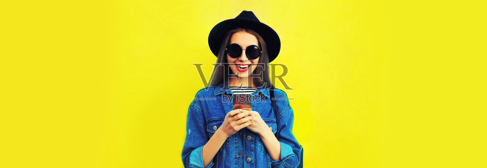 时尚快乐微笑的年轻女性模特的肖像与智能手机戴着黑色圆帽子，牛仔夹克在黄色的背景，空白的拷贝空间的广告文字照片摄影图片