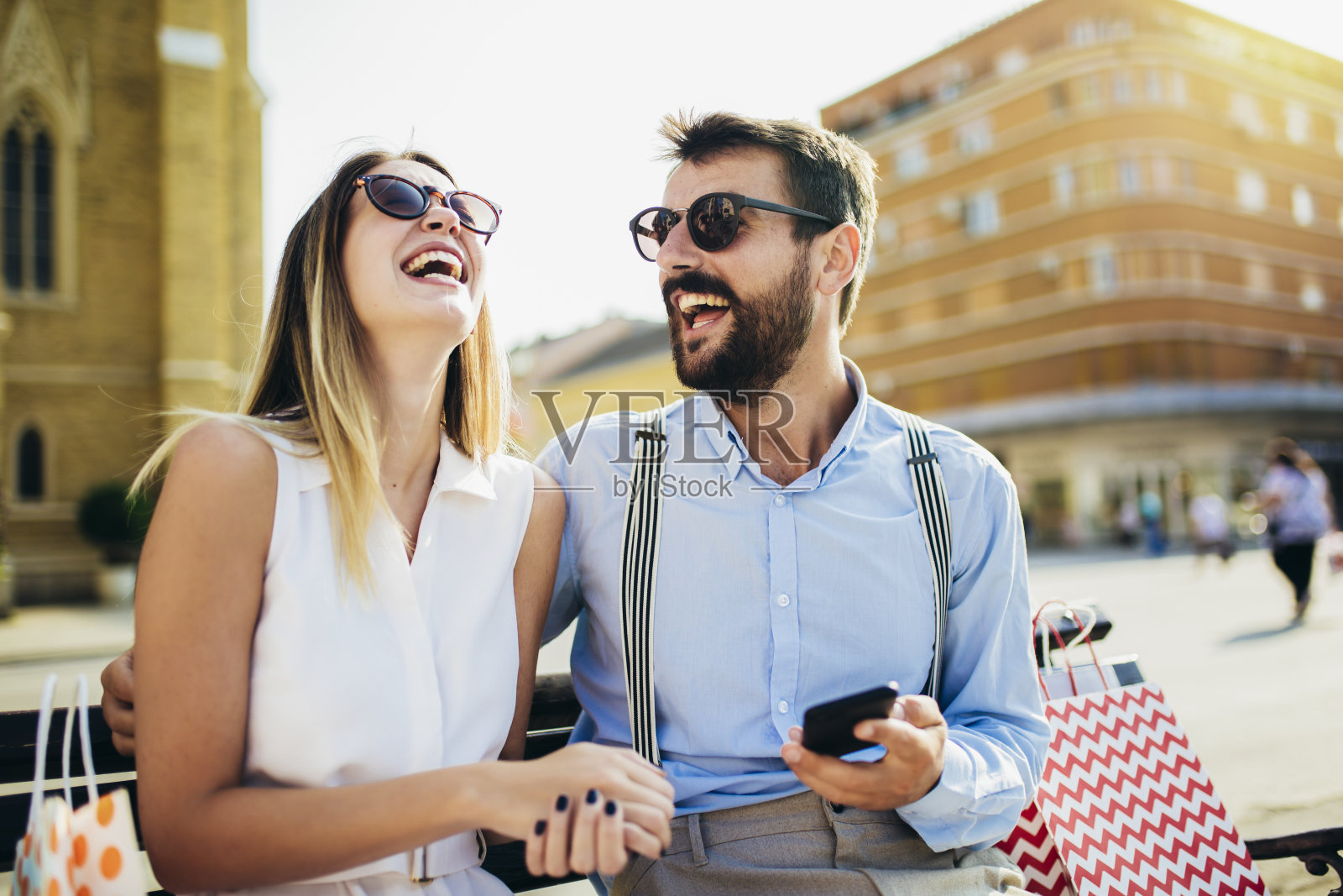 一对年轻的情侣购物后坐在长椅上打电话。照片摄影图片