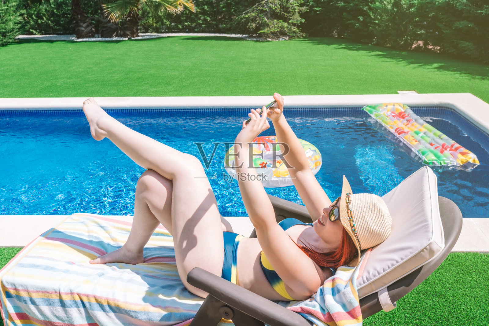 身穿比基尼的年轻女子，在泳池边的阳光躺椅上用她的智能手机自拍。年轻女子在暑假在家晒日光浴。概念夏天和自由时间。照片摄影图片