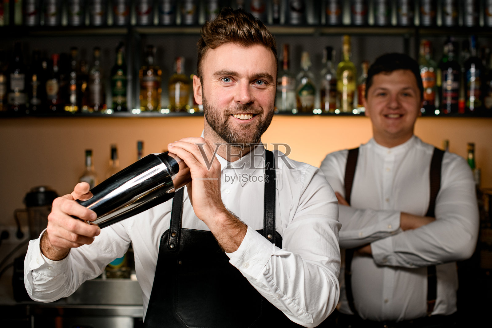 年轻微笑的调酒师在酒吧内部用钢调酒器摇着鸡尾酒照片摄影图片