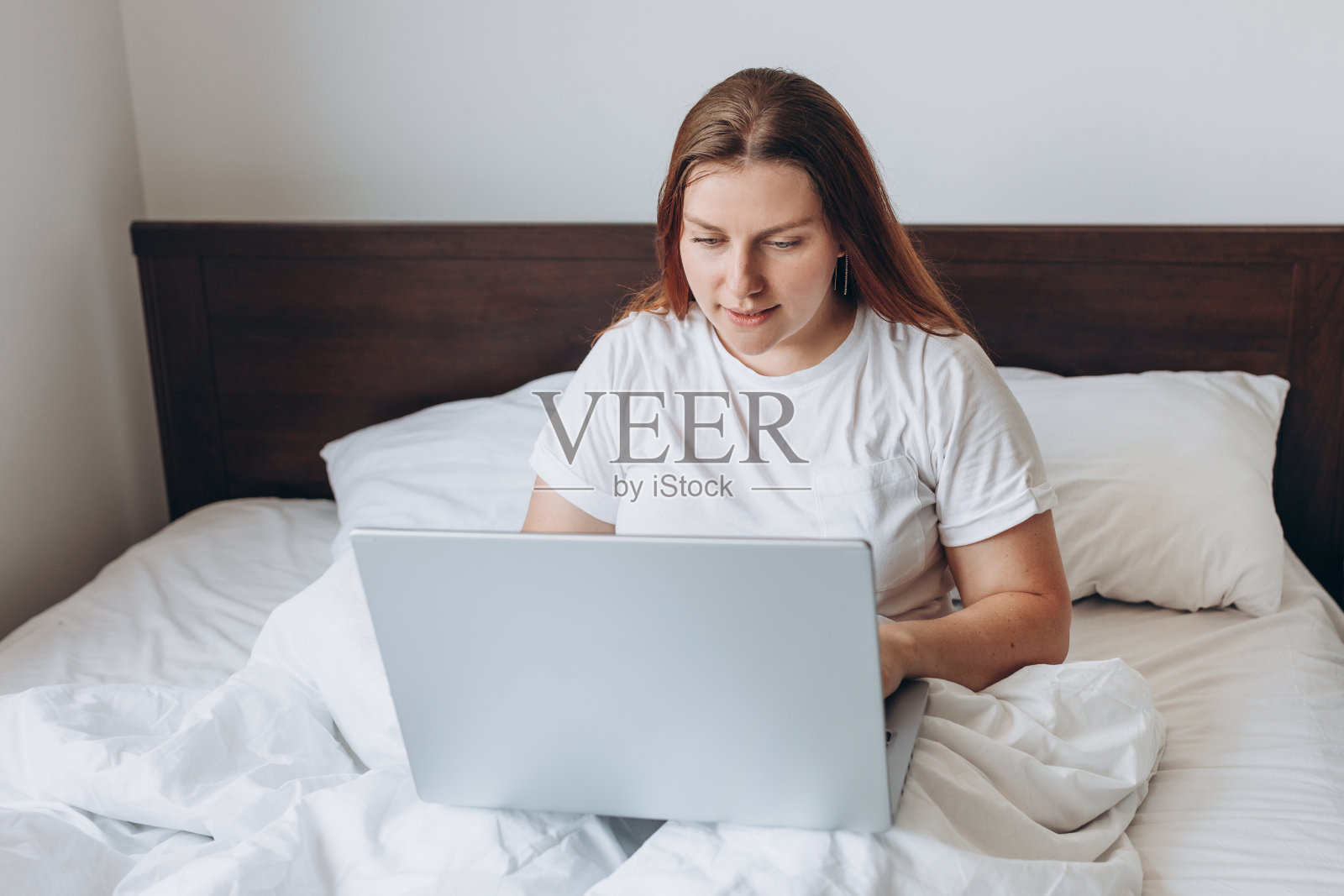 美丽的年轻红发女人的肖像与笔记本电脑在床上懒洋洋的一天。在线教育、工作或购物。替代办公室自由职业者的技术女性概念照片摄影图片