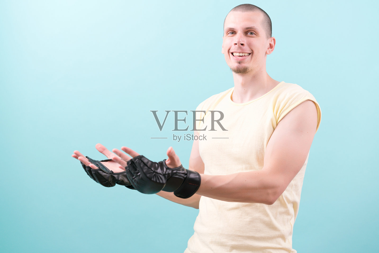 一个快乐的综合格斗选手戴着黑色手套，穿着黄色衬衫站在蓝色的背景上，显示在复制空间上的两只手。自信。冠军。手套。耐力。战斗。战斗。微笑照片摄影图片