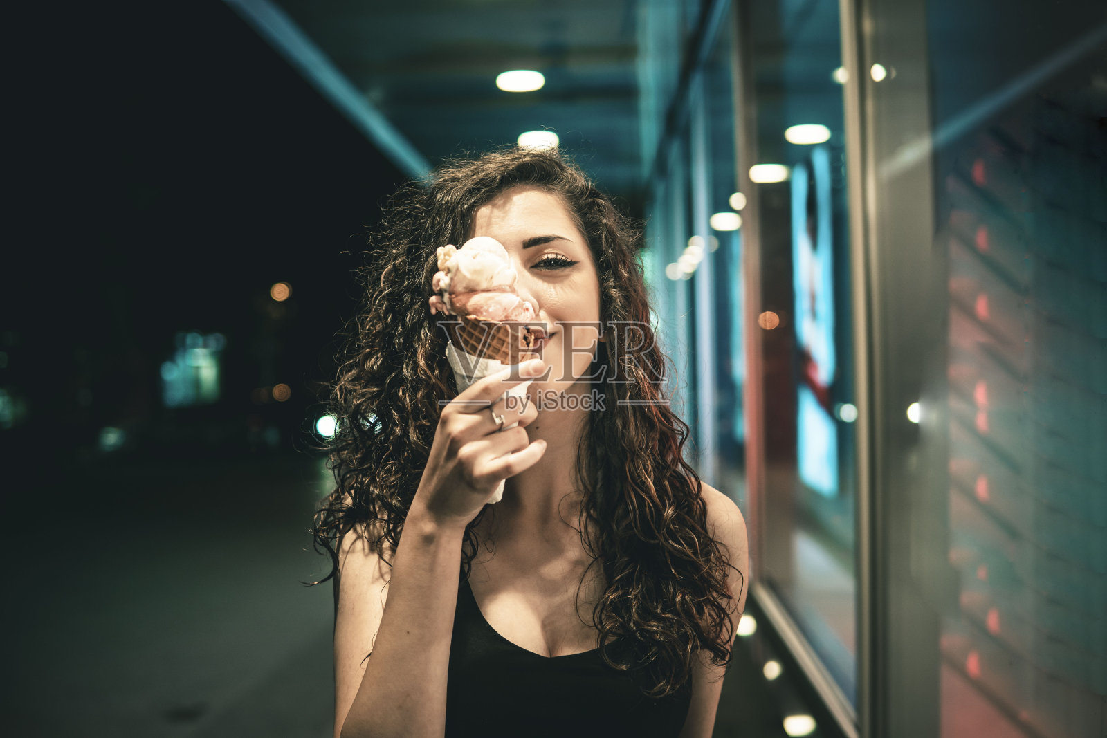 女孩与卷曲的棕色头发持有冰淇淋在一个锥形在她的手覆盖她的半张脸。照片摄影图片