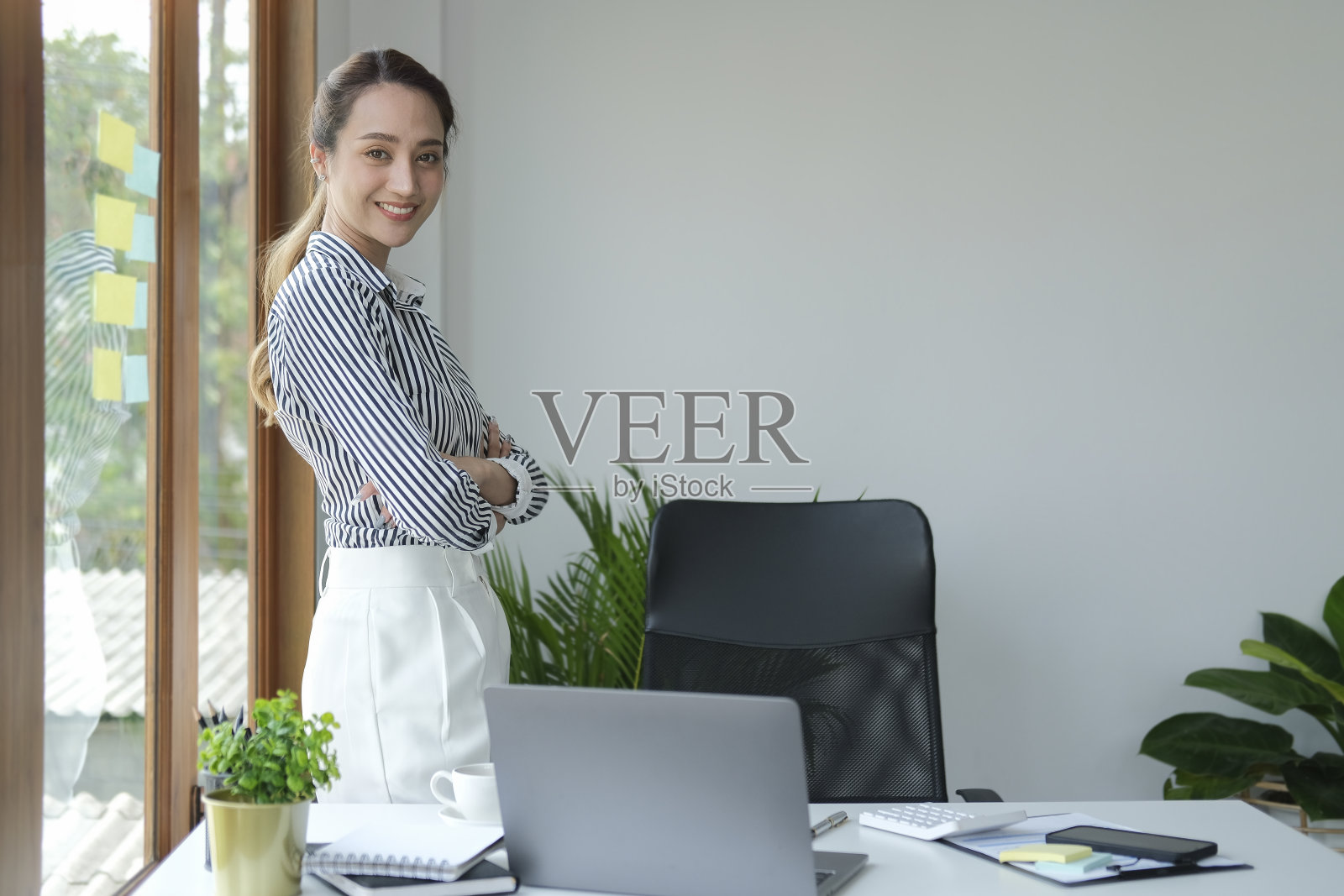 自信的千禧一代女商人双手交叉站在办公室窗户旁，对着镜头微笑。照片摄影图片