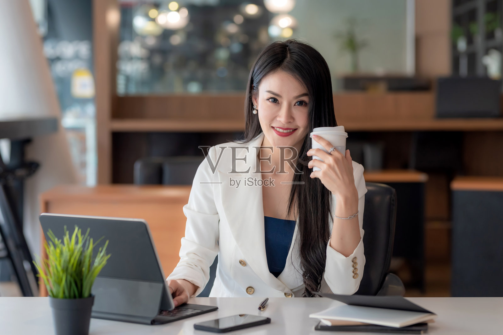 迷人的亚洲女商人工作在办公室拿着咖啡准备在办公室使用平板电脑。看相机。照片摄影图片