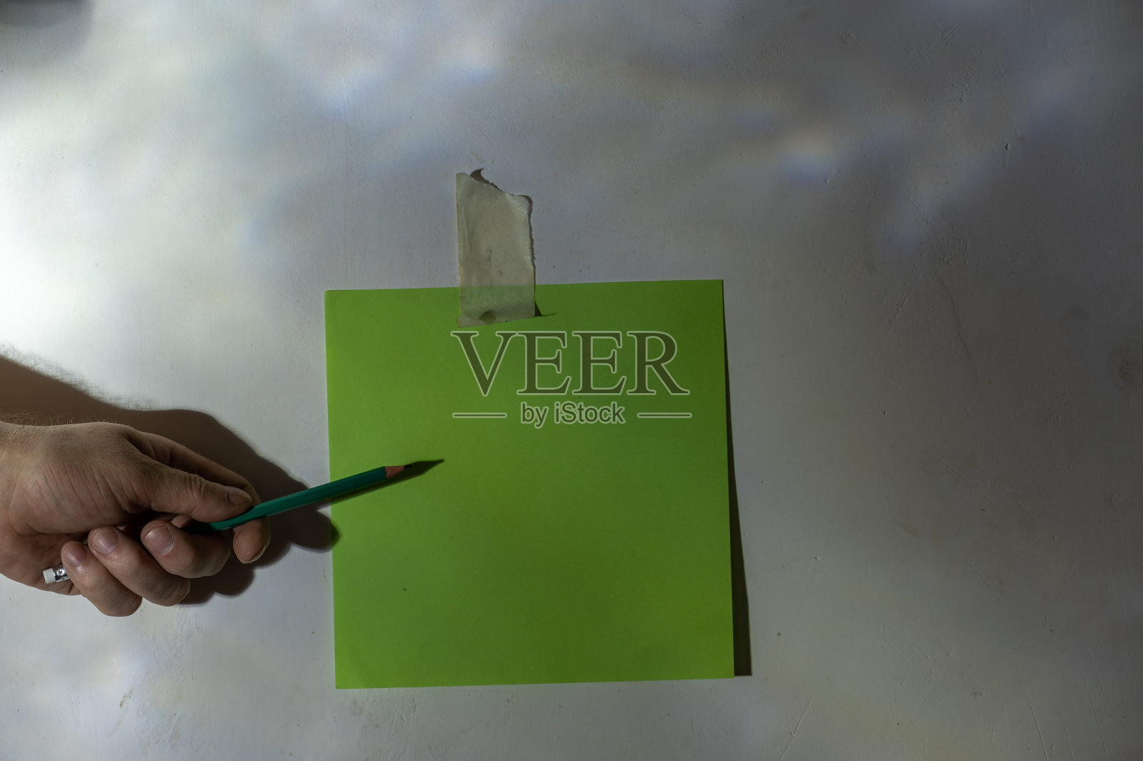 成熟男人的手用铅笔指着一张纸。把一张绿色的方形空白纸固定在墙上。灰色灰泥墙上的抽象光影画。有选择性的重点。低调照片摄影图片