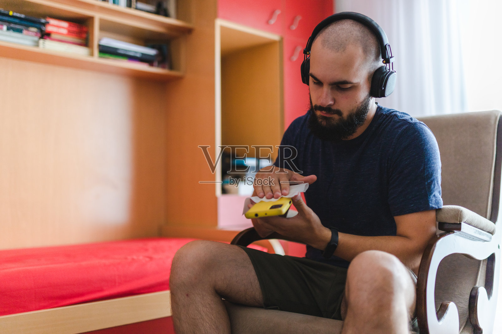 留着胡子的秃顶年轻人在家里一边用湿纸巾擦手机，一边戴着耳机听音乐照片摄影图片