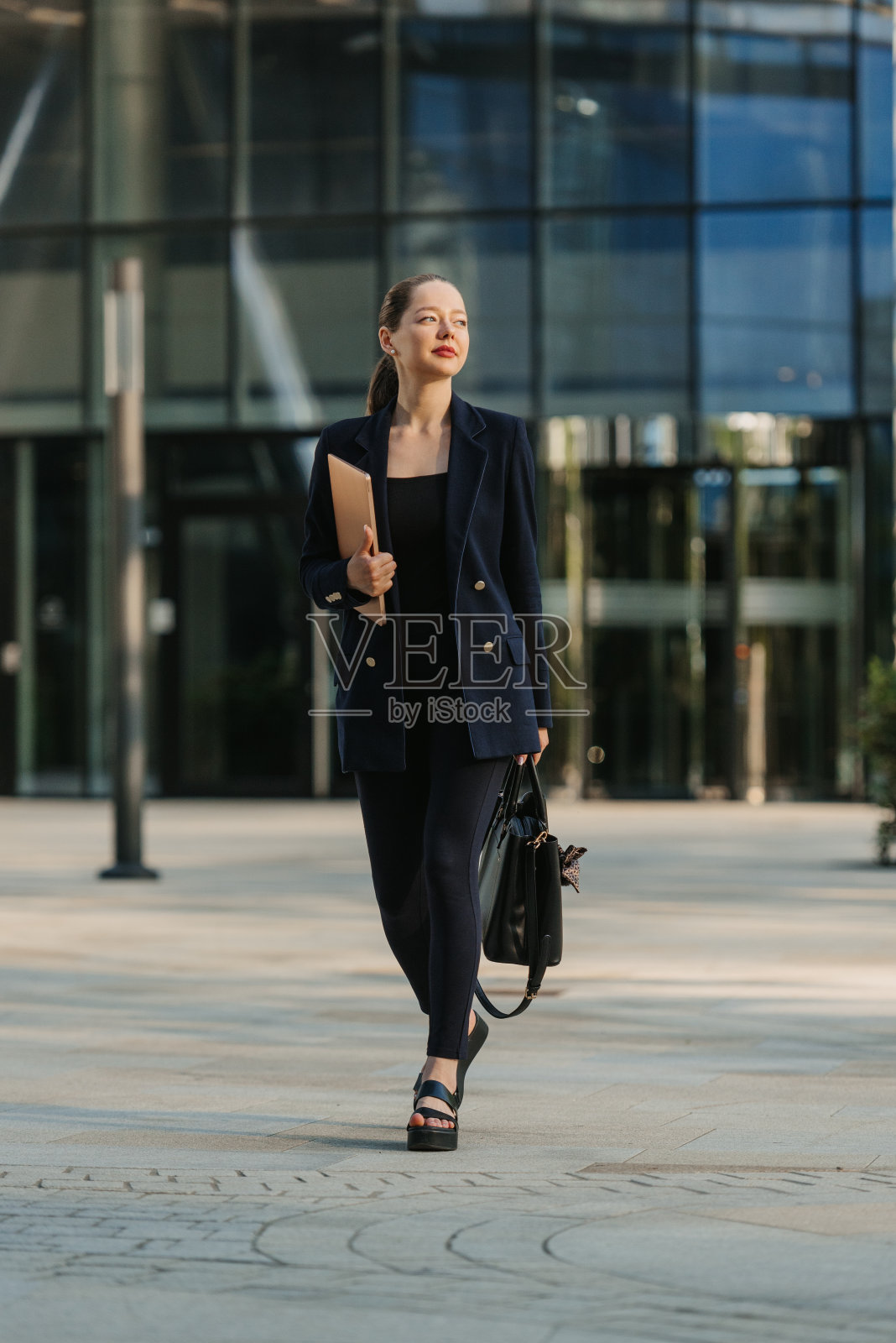 一位穿着西装的女房地产经纪人正拿着一台笔记本电脑在摩天大楼之间穿行。照片摄影图片