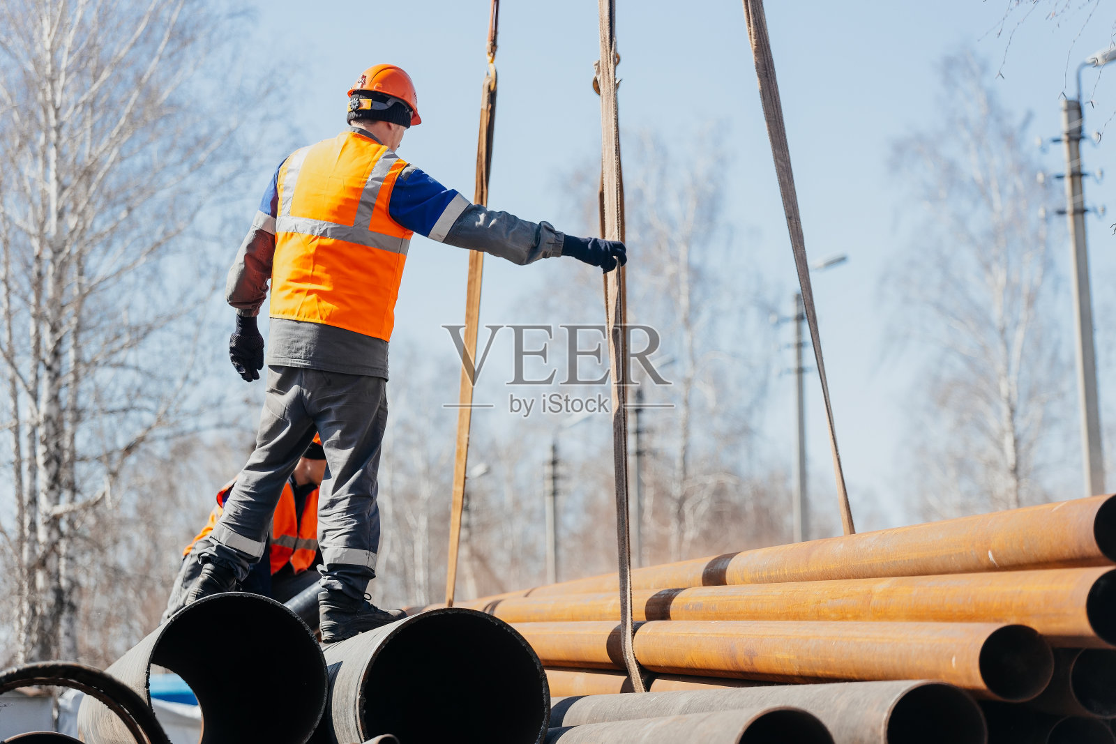 在一个阳光明媚的夏日，戴着头盔的Slinger在工地上卸下金属管道。真实的场景。真正的工人。照片摄影图片