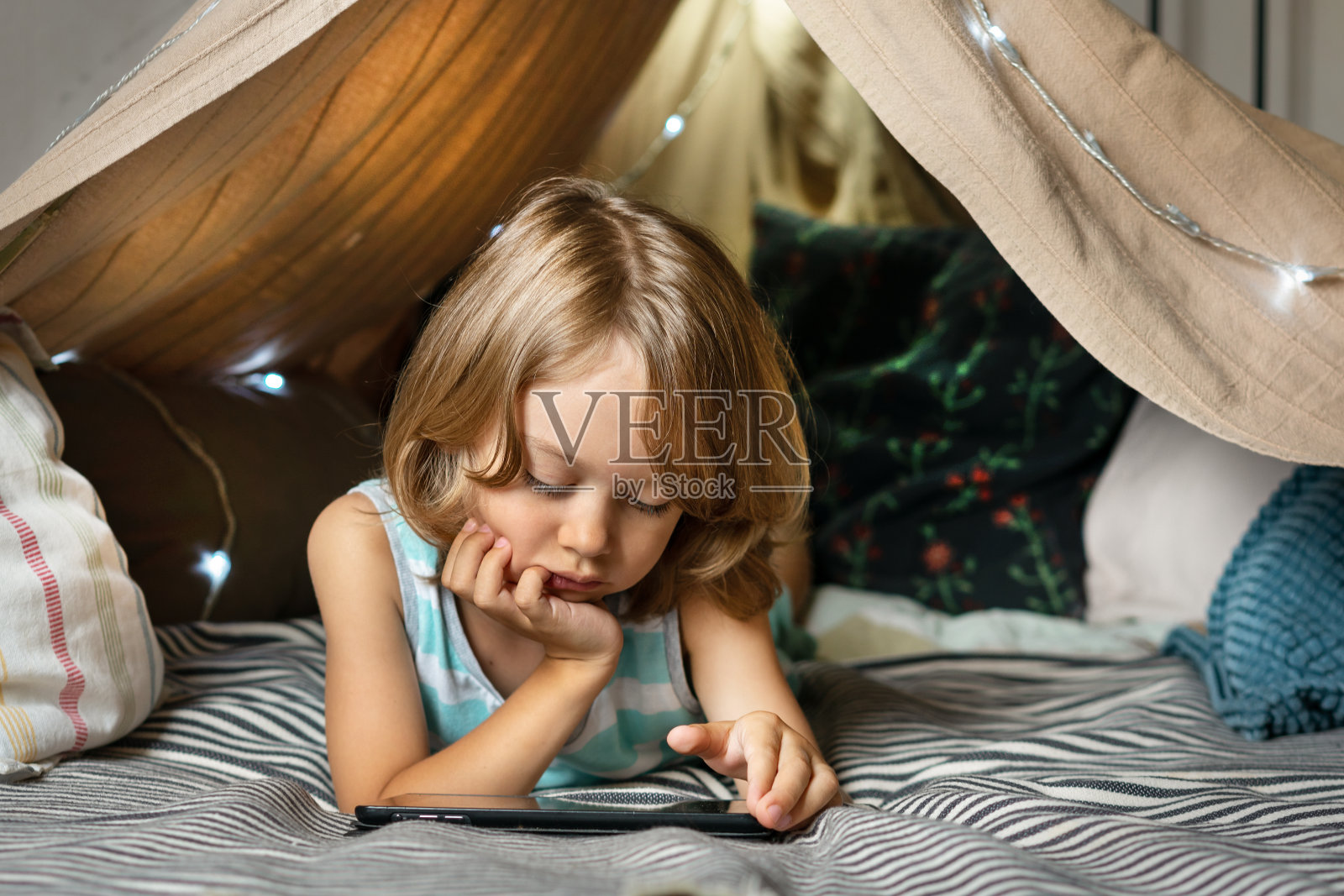 男孩躺在床上用数码平板电脑玩游戏或在家看动画片。照片摄影图片