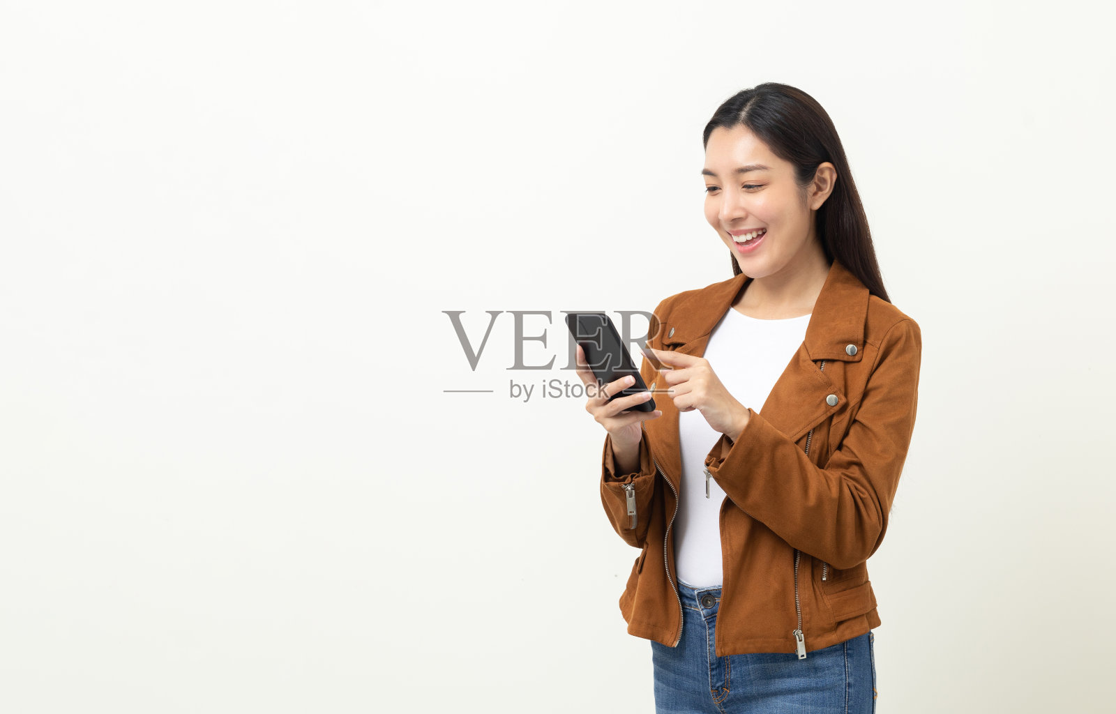 漂亮的年轻亚洲女人使用智能手机站在孤立的白色背景。她用手机发短信。搭配夹克和牛仔裤。照片摄影图片