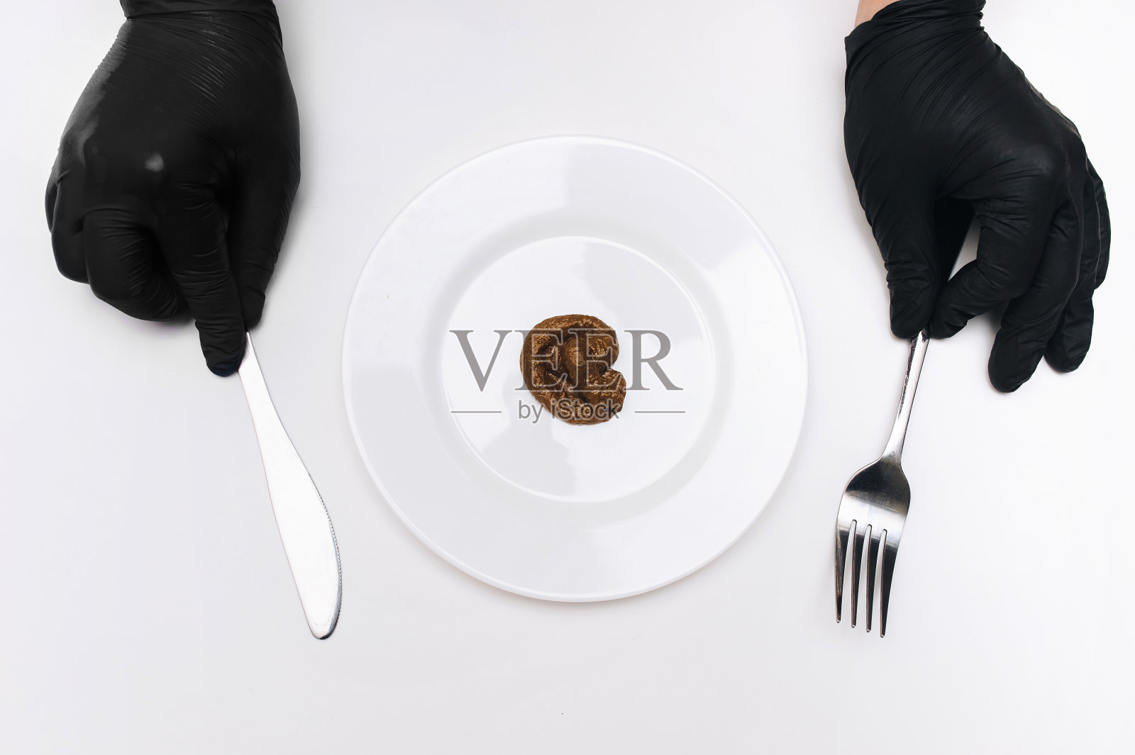 戴着黑色手套的手拿着刀叉，靠近装有人工粪便的白色盘子。假新闻和餐馆劣质食物的概念。照片摄影图片