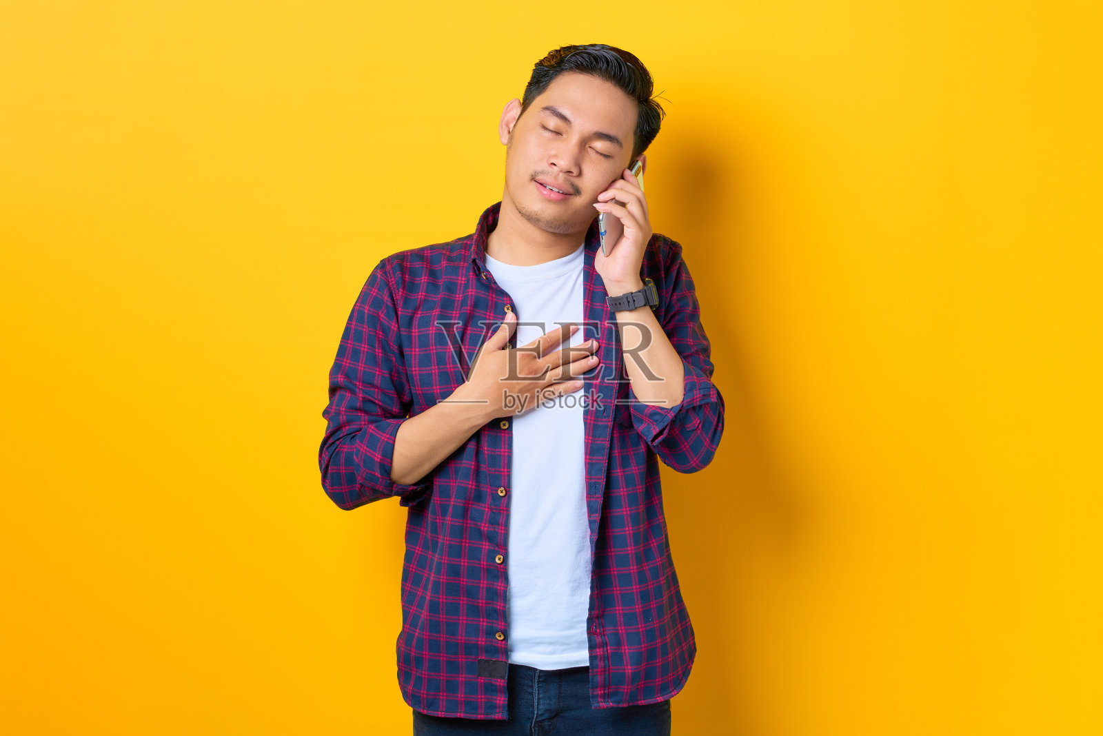 高兴的年轻亚洲男子在格子衬衫谈手机孤立的黄色背景照片摄影图片