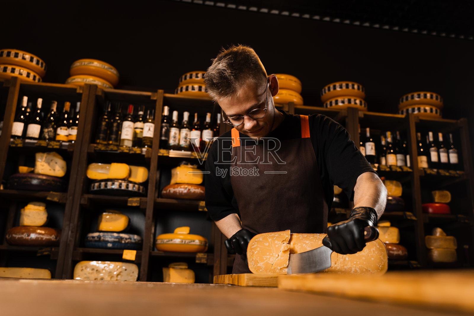 帅气的奶酪侍酒师用刀切限量豪达奶酪。留着胡子戴着时髦眼镜的潮人在奶酪店工作。广告创意。照片摄影图片