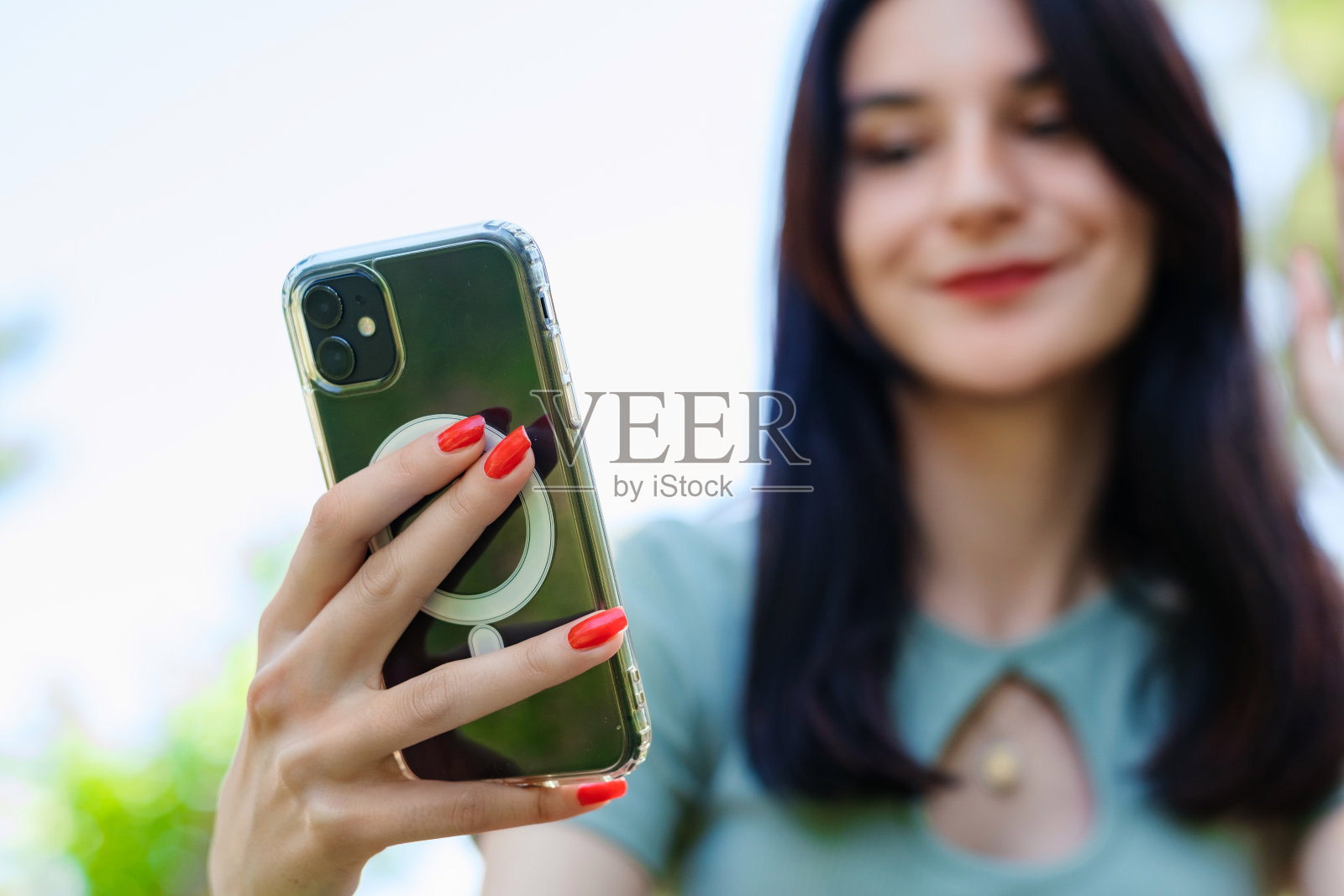 可爱的黑发女子穿着绿松石t恤站在城市公园，在户外拿着手机看屏幕挥手视频呼叫远方的朋友在移动应用程序中使用智能手机视频聊天照片摄影图片