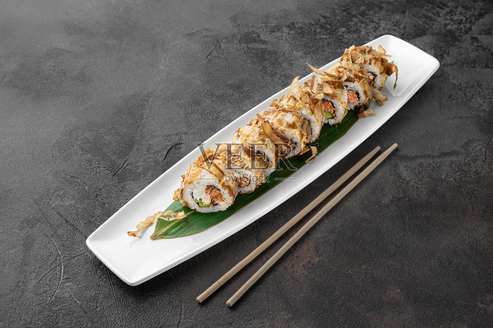 一套鲣鱼卷与烤鲑鱼，鳄梨，金枪鱼片和绿色竹叶在一个白色陶瓷盘子与筷子在深灰色的纹理背景，侧视图照片摄影图片