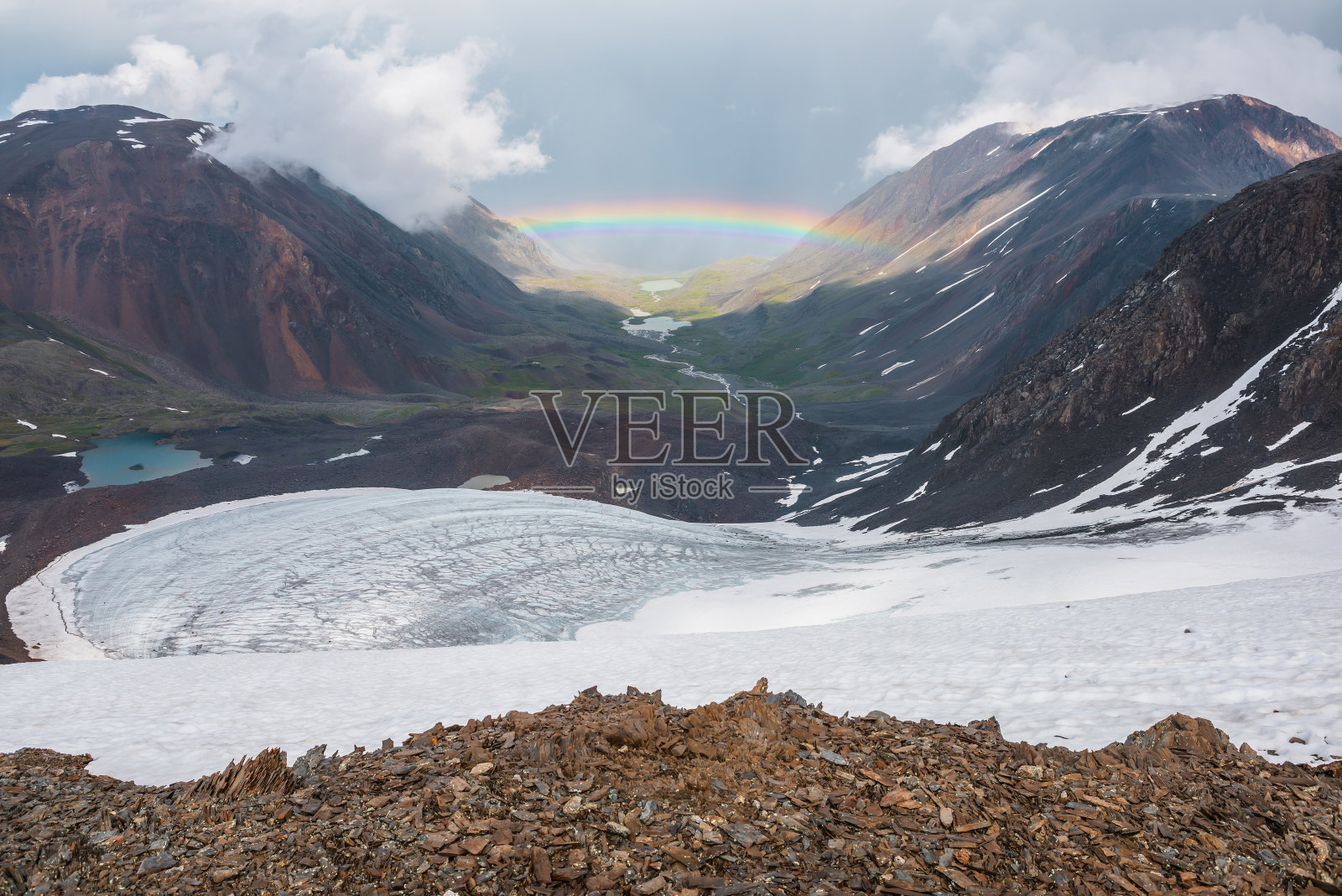 荒凉的风景和生动的彩虹上方的山湖。阴郁的风景与明亮的彩虹上方的冰川和冰川湖在山谷。山顶上五颜六色的彩虹和低云。照片摄影图片