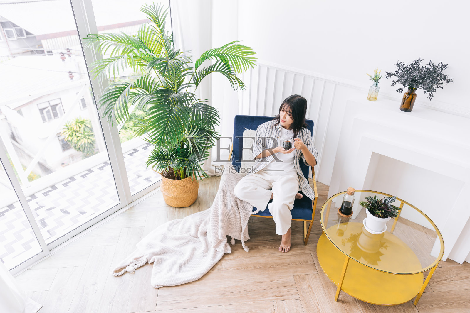 年轻漂亮的女人微笑着坐在现代生活区的椅子上，绿色的植物，放松地喝着一杯热咖啡，在一个舒适的家的室内环境中治愈倒影。照片摄影图片