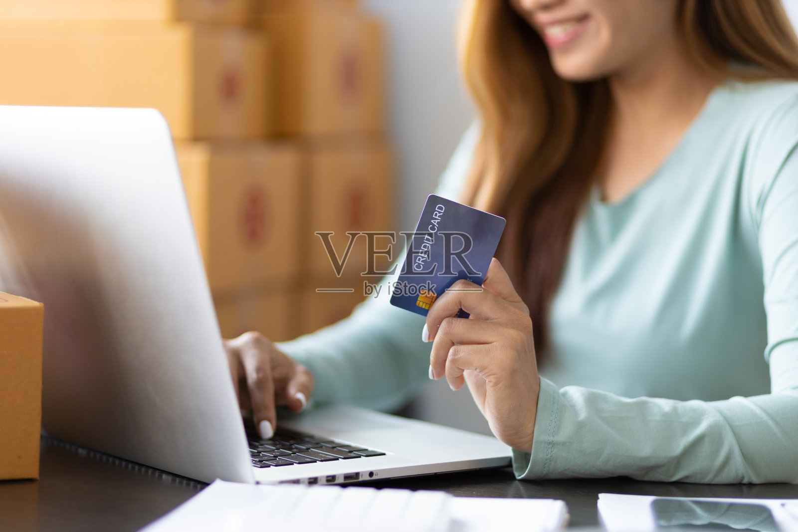 中小企业经营者正在用笔记本电脑检查网上订单的余额。小型企业的企业家持有信用卡，用笔记本电脑和家庭办公室的盒子工作。网上购物的女性。照片摄影图片