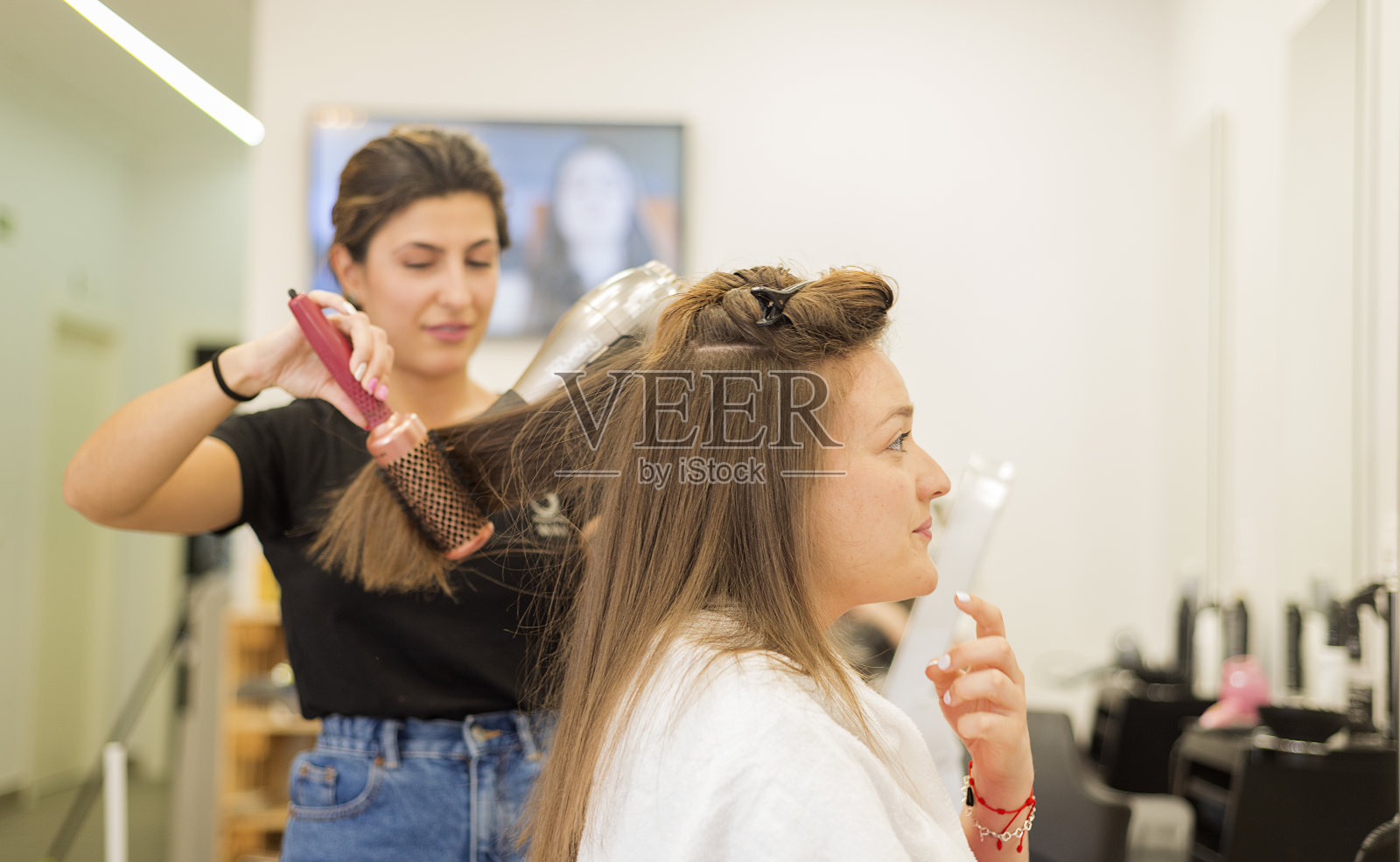 微笑的年轻女子，她的头发在理发店用吹风机吹干。照片摄影图片