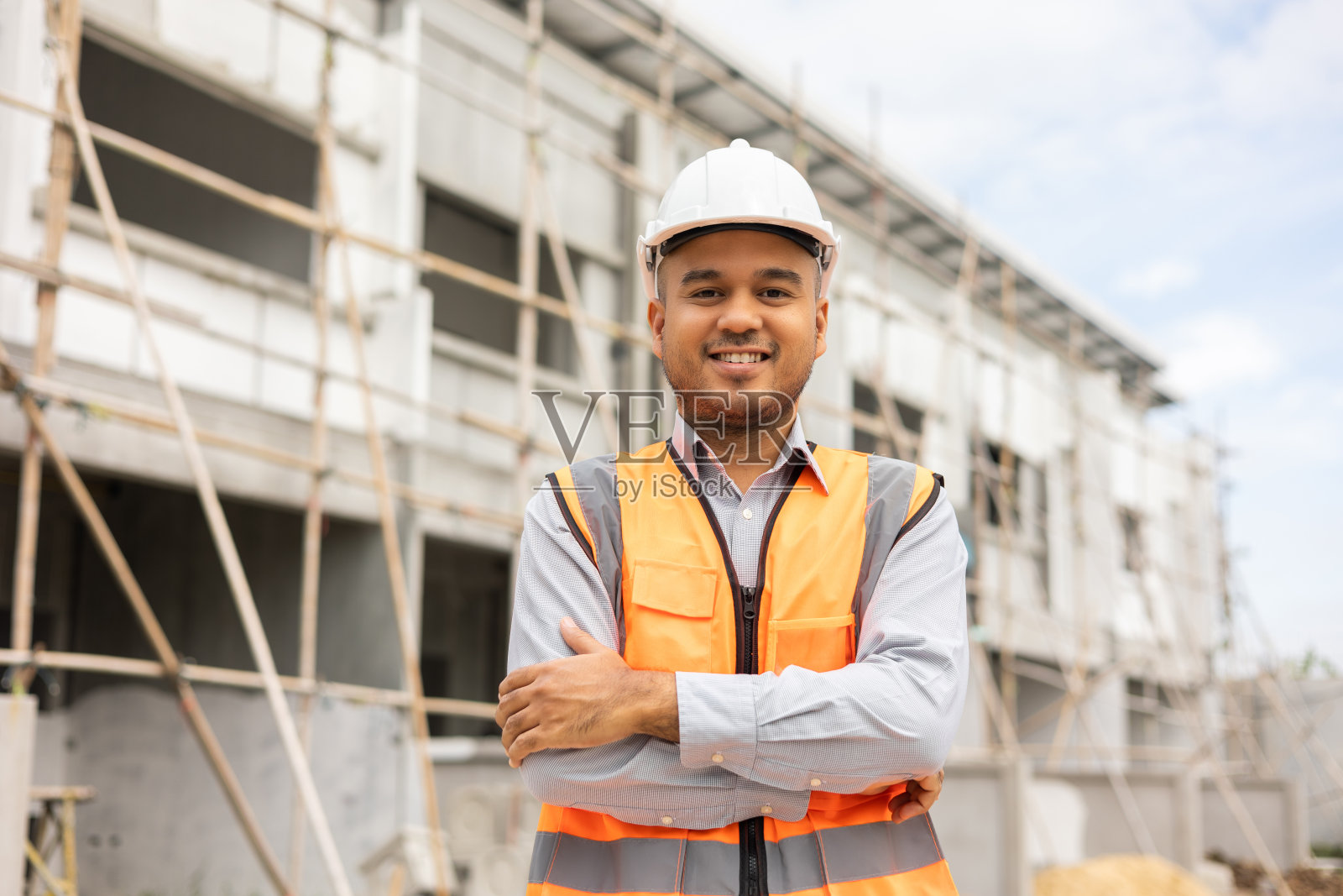 自信的工程师英俊男子双臂交叉站在现代住宅建筑施工。建筑工地上戴白色安全帽的建筑师。照片摄影图片