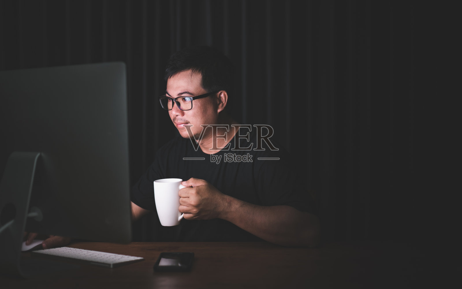 早起概念:亚洲人拿着白色咖啡杯，在黑暗的房间里对着电脑工作照片摄影图片