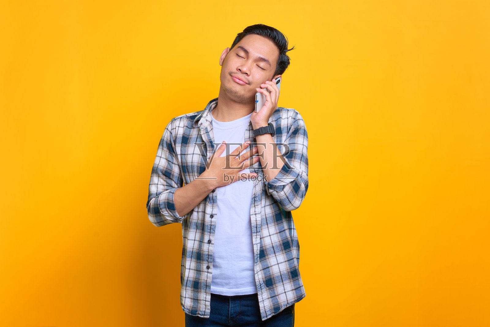 高兴的年轻亚洲男子在格子衬衫谈手机孤立的黄色背景照片摄影图片