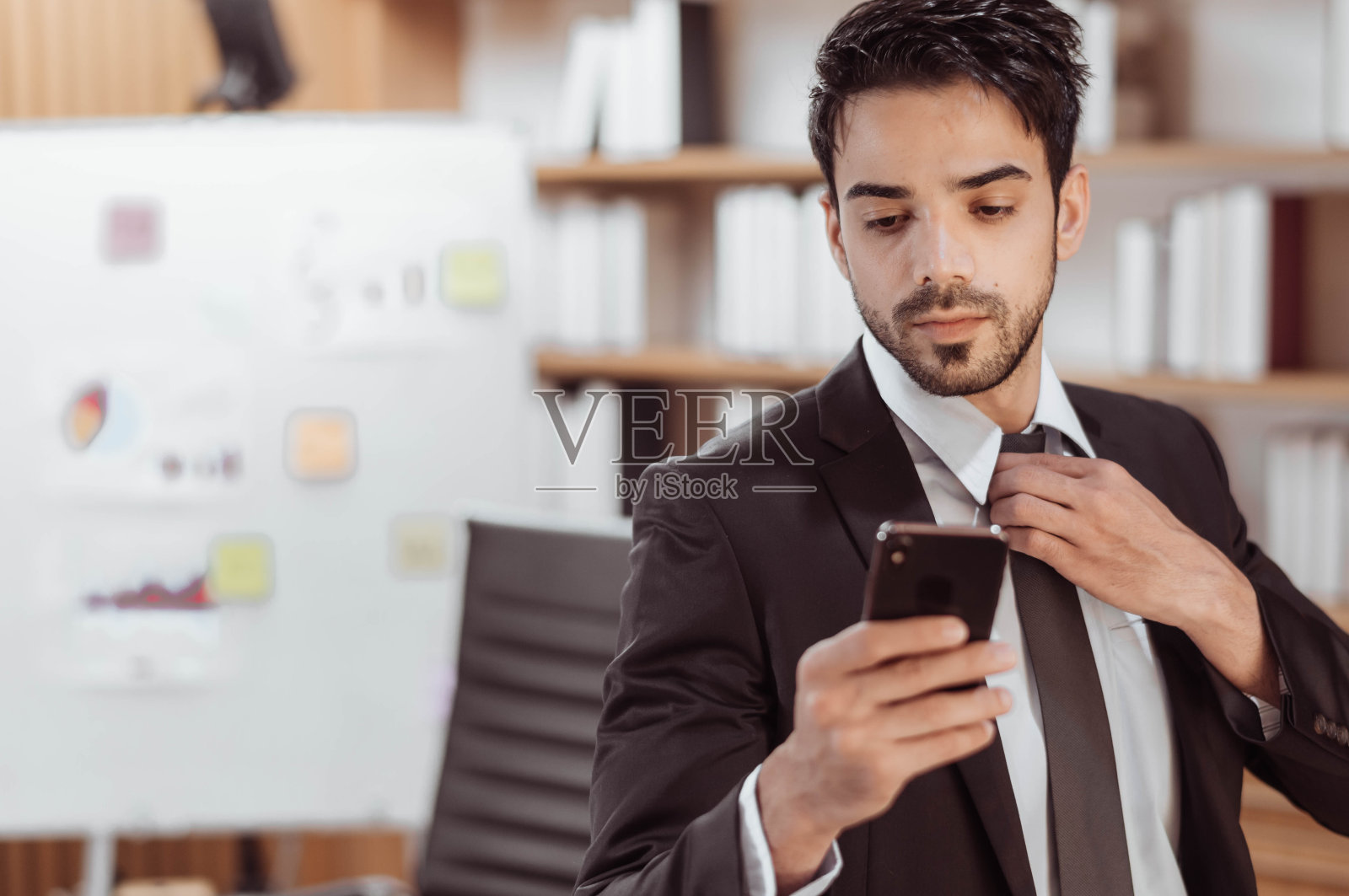 自信的商人在工作室内使用智能手机的画像。英俊的阿拉伯商人在黑色西装和手机在办公室。照片摄影图片